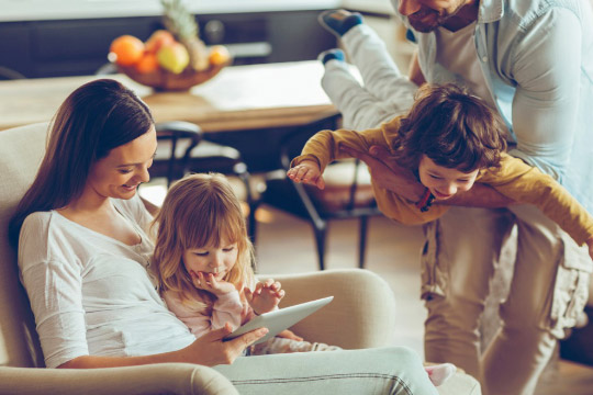 Eine Mutter sitzt mit zwei Kindern auf einem Sofa und schaut auf ein Tablet, während ein Vater einen der Kinder trägt.