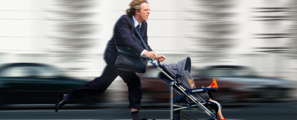 Ein Mann in Anzug eilt mit einem Kinderwagen durch die Stadt, während Autos im Hintergrund vorbeifahren.