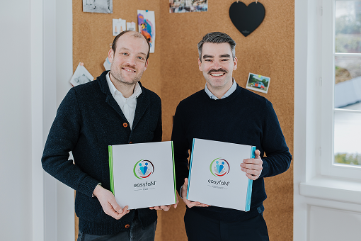 Two men each hold a box with the "easyfam" logo, in front of a cork board with photos.