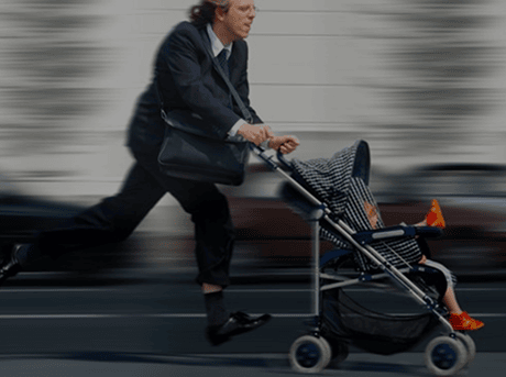 Ein Mann im Anzug rennt mit einem Kinderwagen auf der Straße. Ein Kind sitzt darin, während er hastig unterwegs ist.